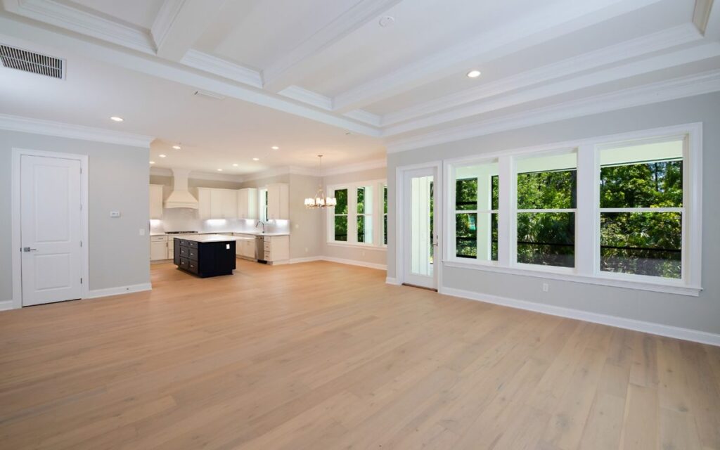 Bright open-concept living area with light wood floors, white cabinetry, and a kitchen island; large windows overlook greenery.