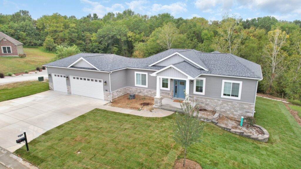 Modern gray one-story home with a three-car garage and stone foundation, front porch, and well-kept lawn.0b1 Shade-roofed house with white trim set in a suburban yard near trees, aerial view.