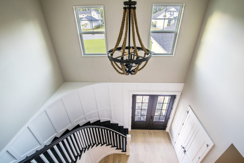 Curved staircase with black railing, white paneled walls, and a rope chandelier hanging from the ceiling in a bright foyer.